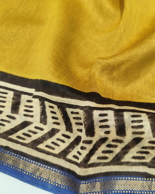 A close-up of a yellow Maheshwari Silk Cotton saree, featuring a wide decorative border with black geometric patterns, beige details, and a blue and gold zari trim.