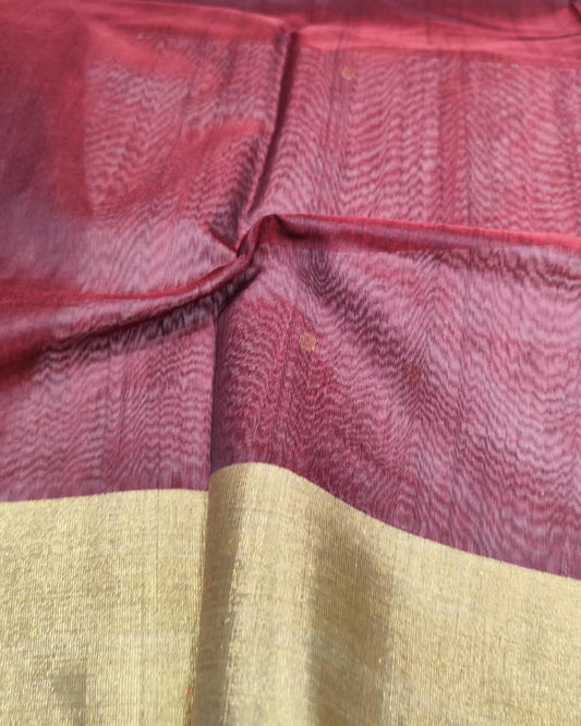 A close-up of a draped fabric, likely a saree, featuring a deep maroon body with a subtle wavy pattern and a wide, shimmering gold border at the bottom.