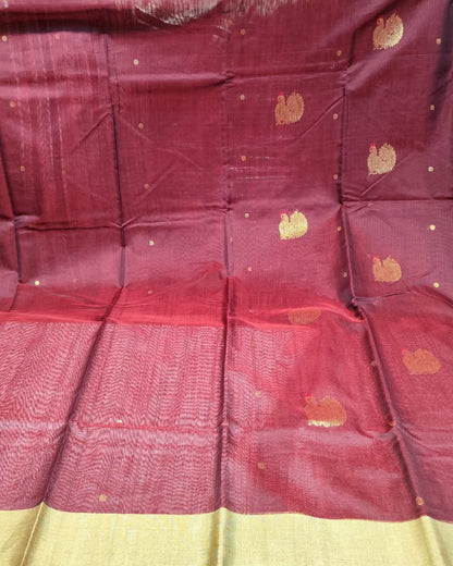 A deep maroon red silk saree with a wide beige border and scattered gold motifs, displayed in a folded and draped layout with a stack of other folded fabrics visible in the background.