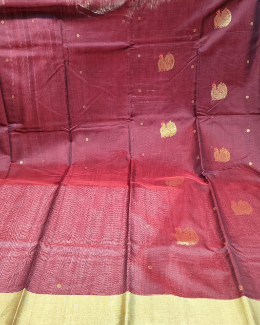 A deep maroon red silk saree with a wide beige border and scattered gold motifs, displayed in a folded and draped layout with a stack of other folded fabrics visible in the background.