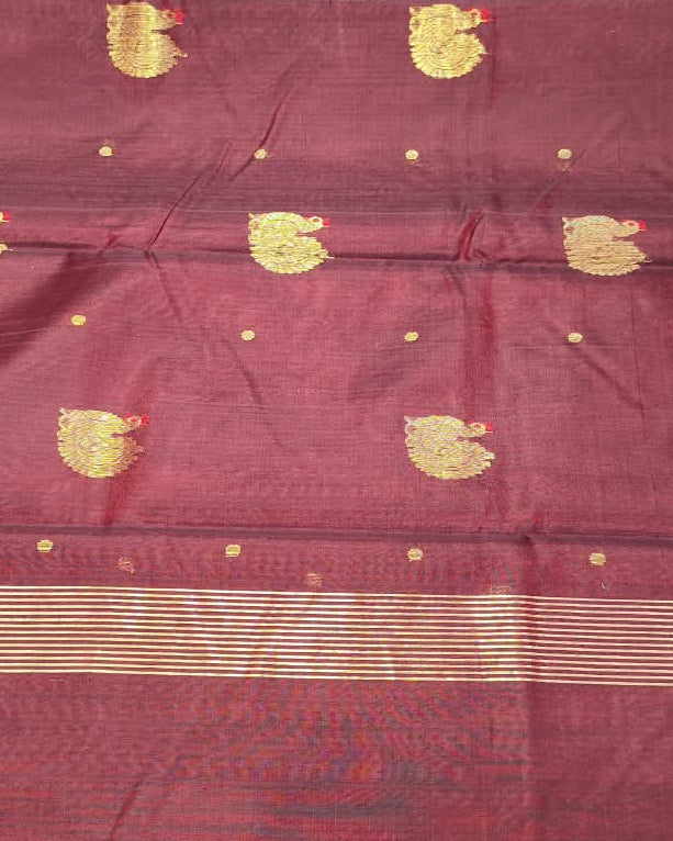 A maroon red silk saree with gold zari motifs and a gold border, held up by a person in a fabric store.