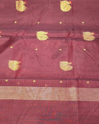 A maroon red silk saree with gold zari motifs and a gold border, held up by a person in a fabric store.