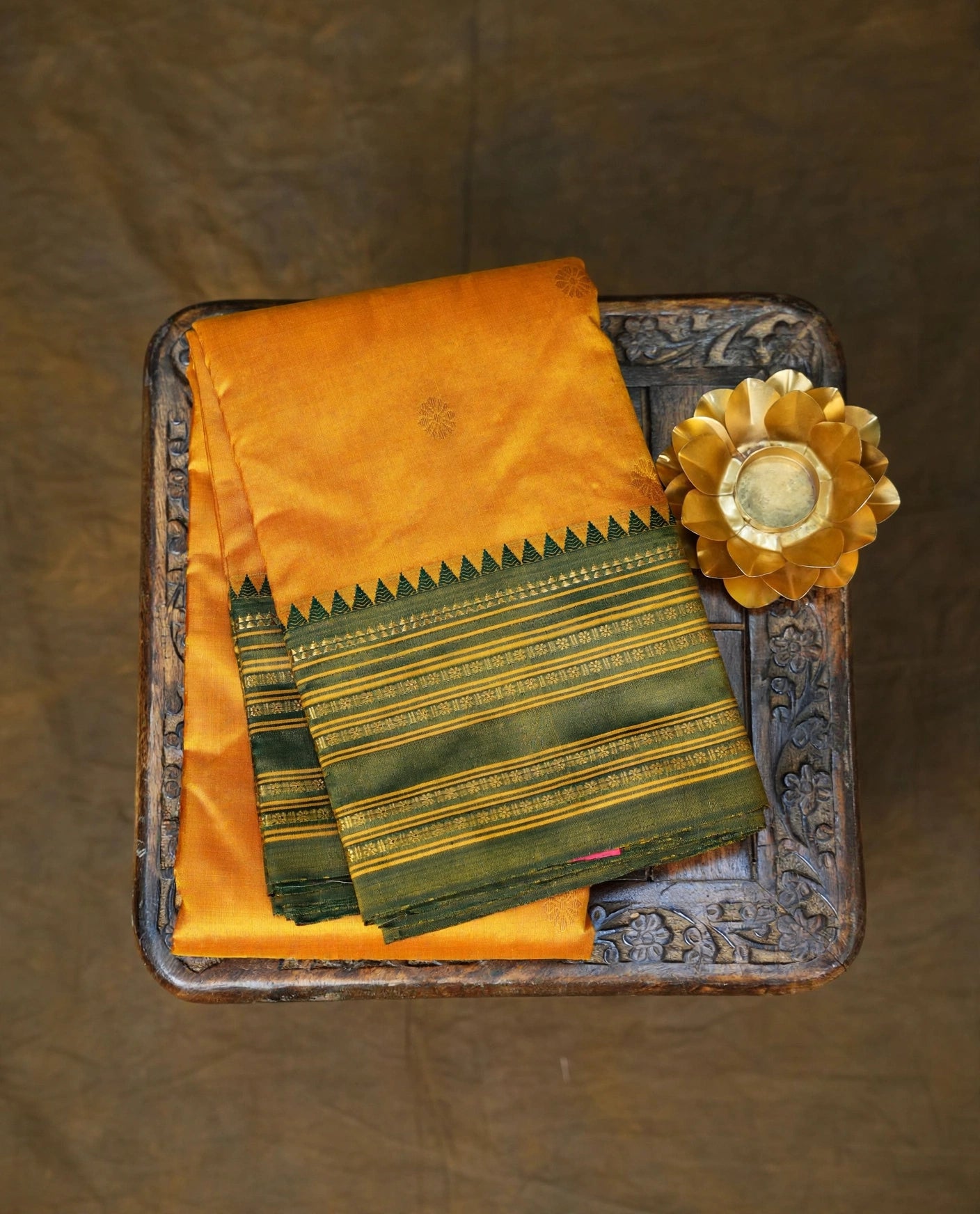 A folded yellow and green silk fabric, likely a saree or dupatta, displayed on an ornate carved dark wooden tray with a gold lotus prop, set against a textured brown backdrop.