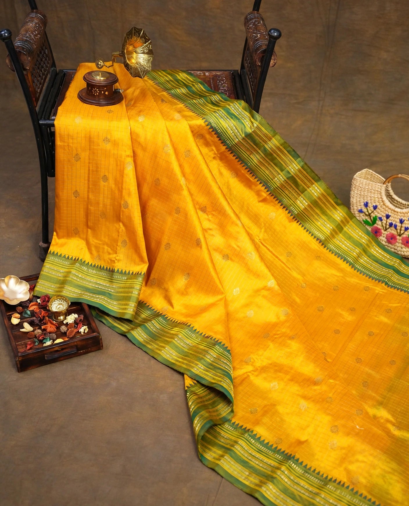 A bright yellow silk saree with green and gold striped borders, draped elegantly over a dark wooden chair in a styled studio setting.