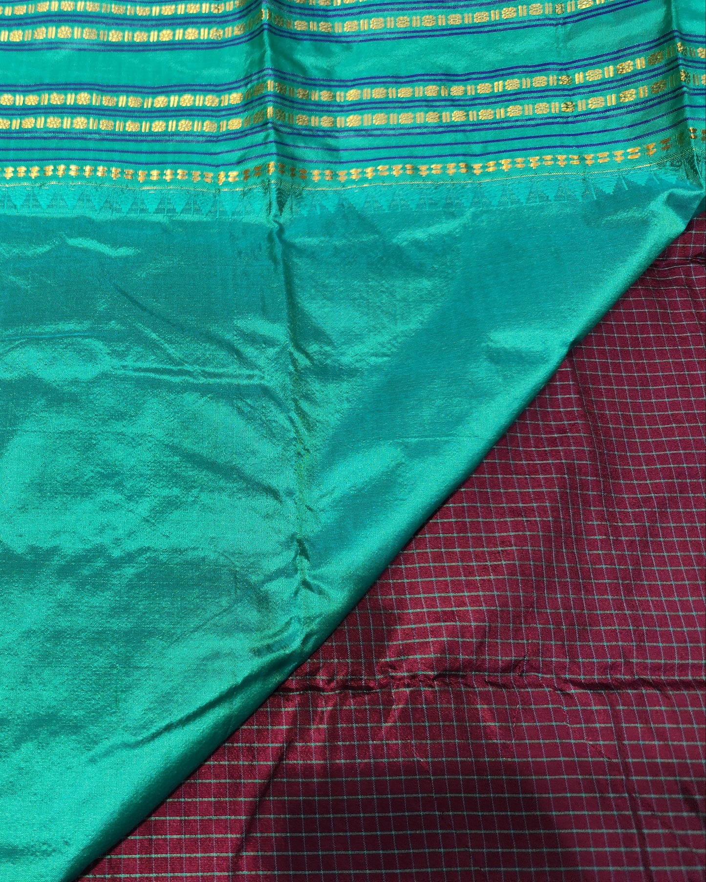 A close-up of a teal greensari, showing its sheen and texture. The fabric features horizontal rows of gold and yellow woven metallic patterns along the border.