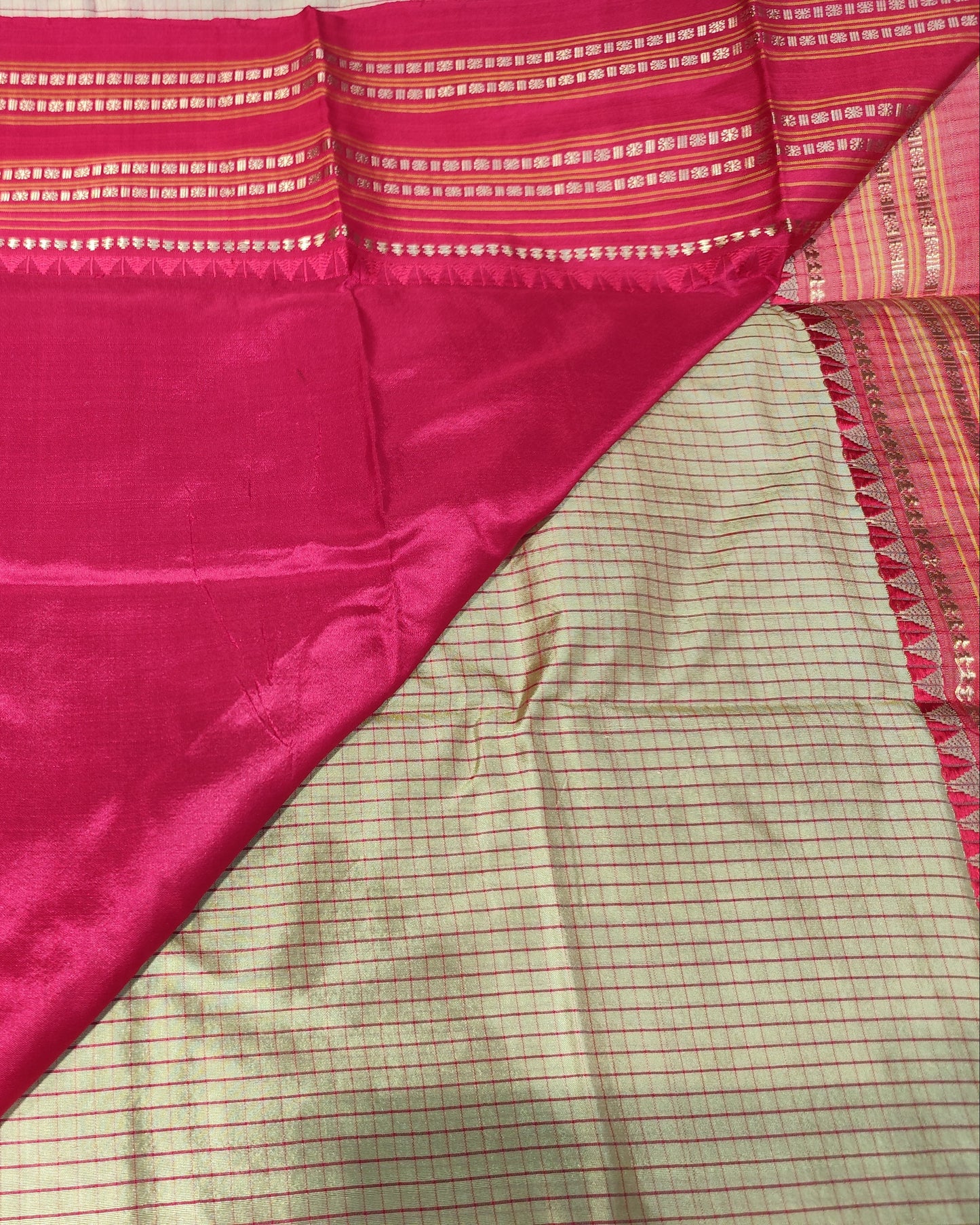 A close-up of a folded and draped fabric, likely a sari, showing a rich magenta-pink body with a woven border, alongside a beige and pink checkered patterned section.
