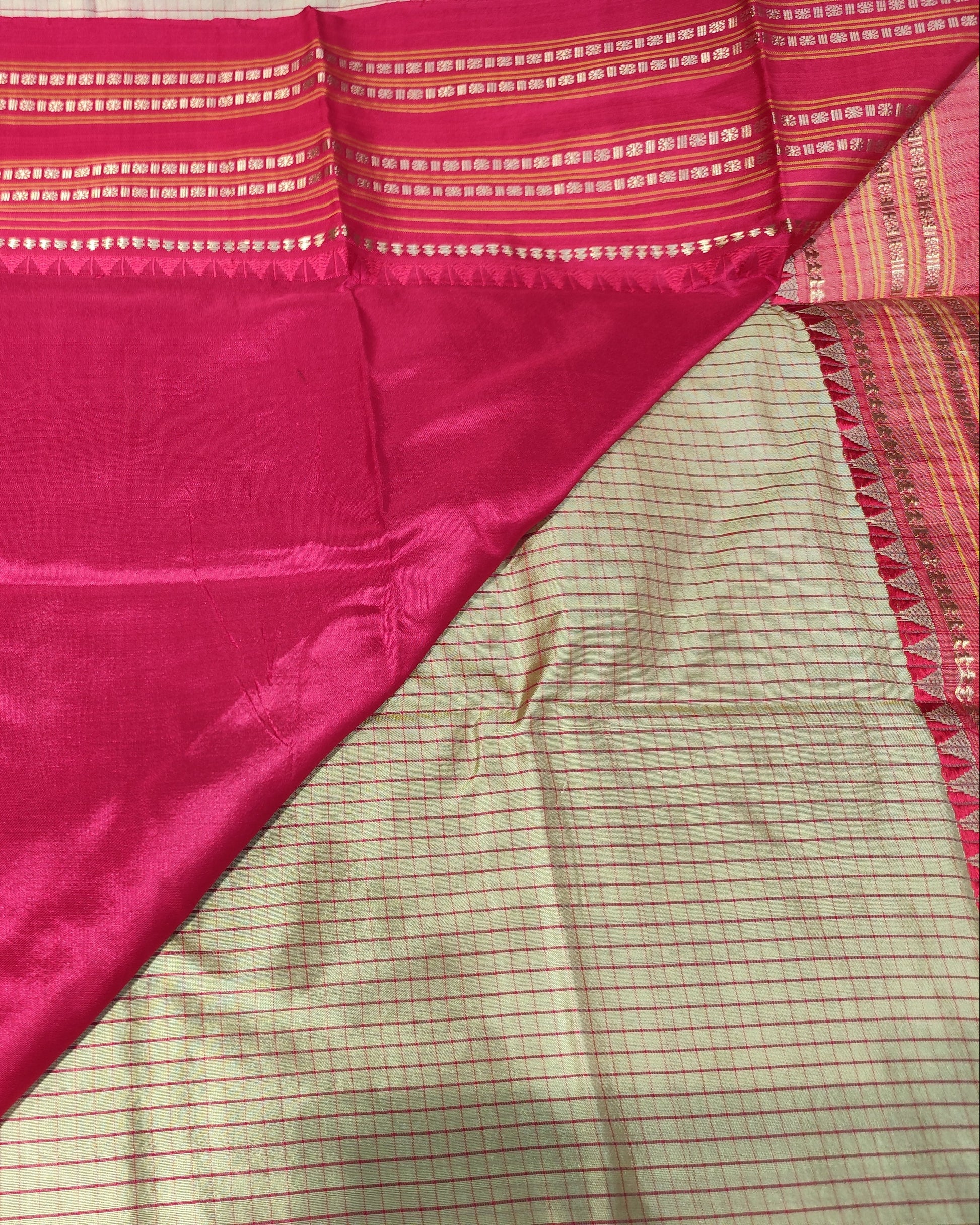 A close-up of a folded and draped fabric, likely a sari, showing a rich magenta-pink body with a woven border, alongside a beige and pink checkered patterned section.