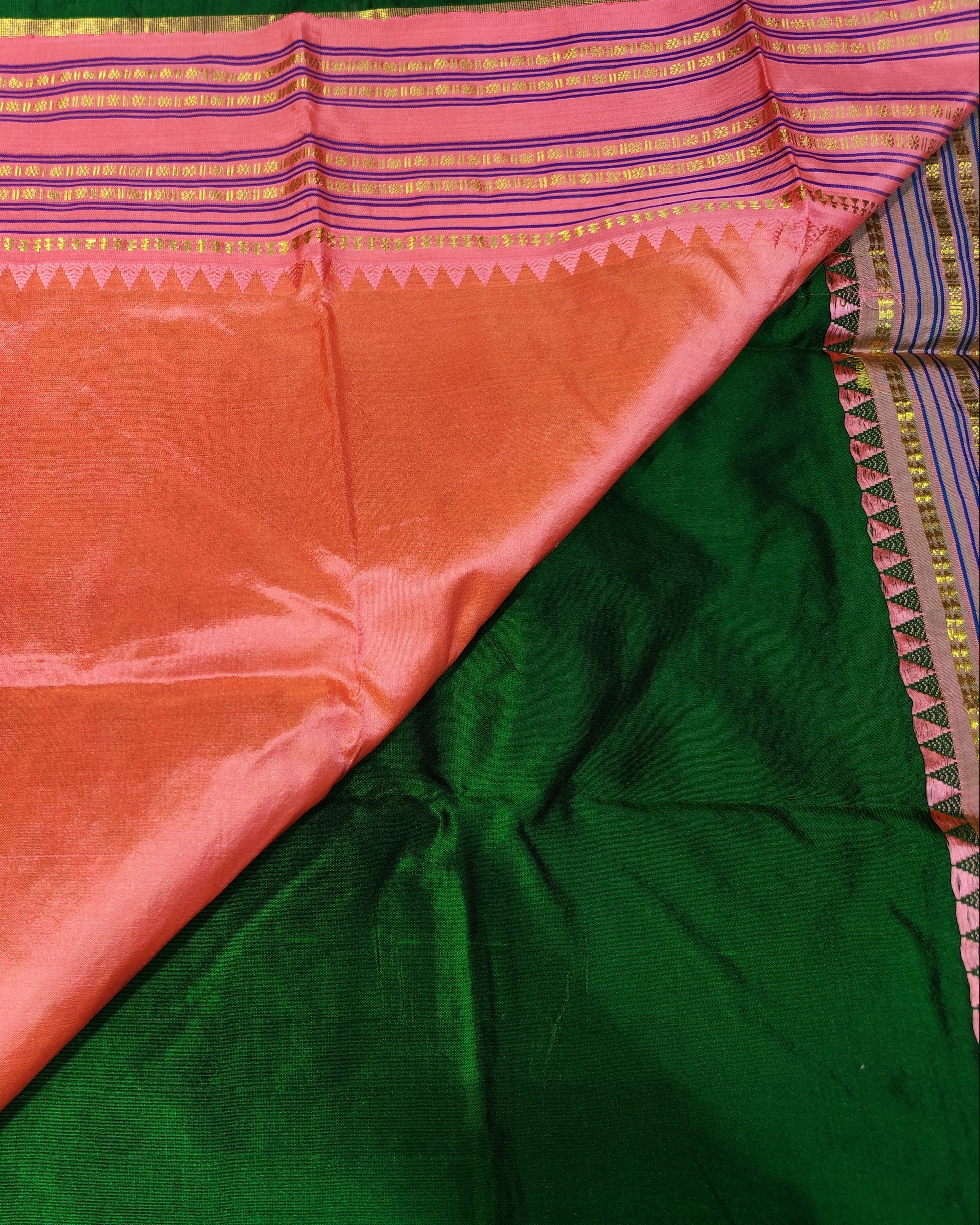 A close-up of a folded and draped silk saree fabric, showing a dominant peach-pink body, a contrasting dark green section, and a decorative border with pink, purple, and gold stripes.