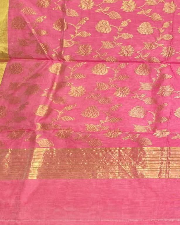 A pink silk saree with gold floral motifs and a wide gold border, held up by a person in a fabric store.