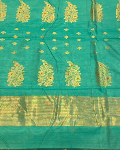 A turquoise blue and gold woven fabric, likely a saree or scarf, held up by a person in a shop setting with other folded textiles in the background.