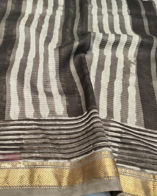 A close-up of a striped Maheshwari Silk Cotton saree, featuring vertical stripes in dark brown, gray, and off-white, with a prominent gold zari border.