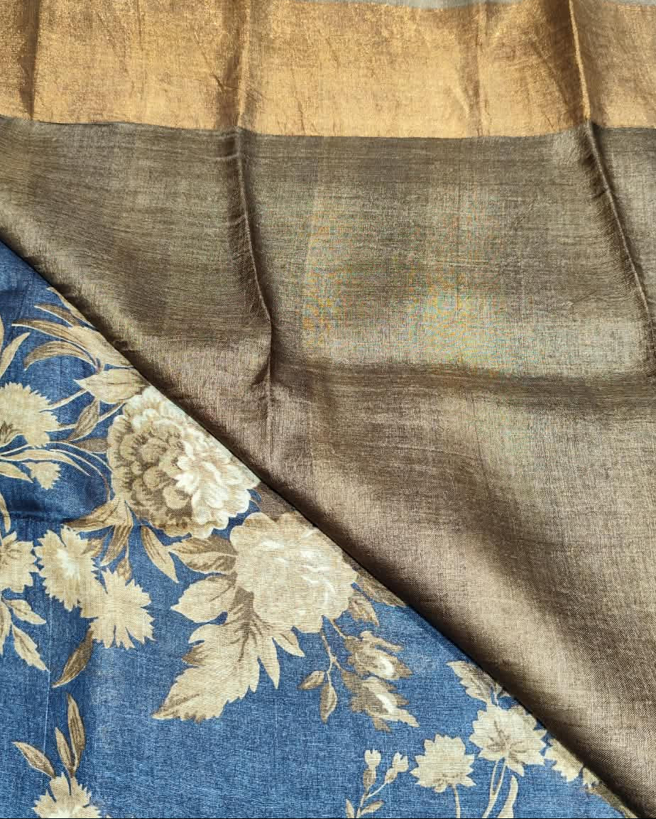 A close-up of a Tussar Silk sari, showing a large taupe-brown field with a wide gold border, and a blue floral section with cream flowers and green stems.