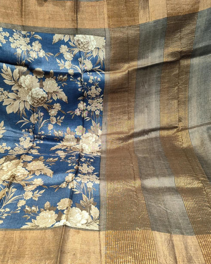 A close-up of a blue and beige floral Tussar silk saree  showing the detailed floral pattern on the body and the striped gold and gray border.