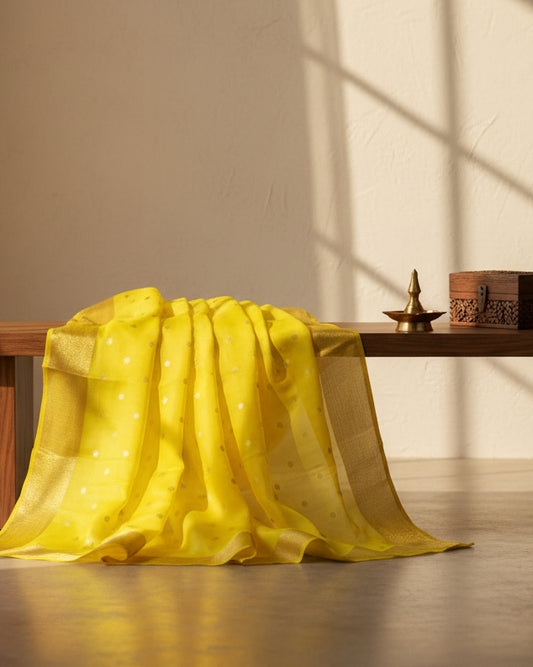A close-up of a bright yellow-green fabric, likely a saree, featuring small metallic gold polka dots and a wide gold zari border.