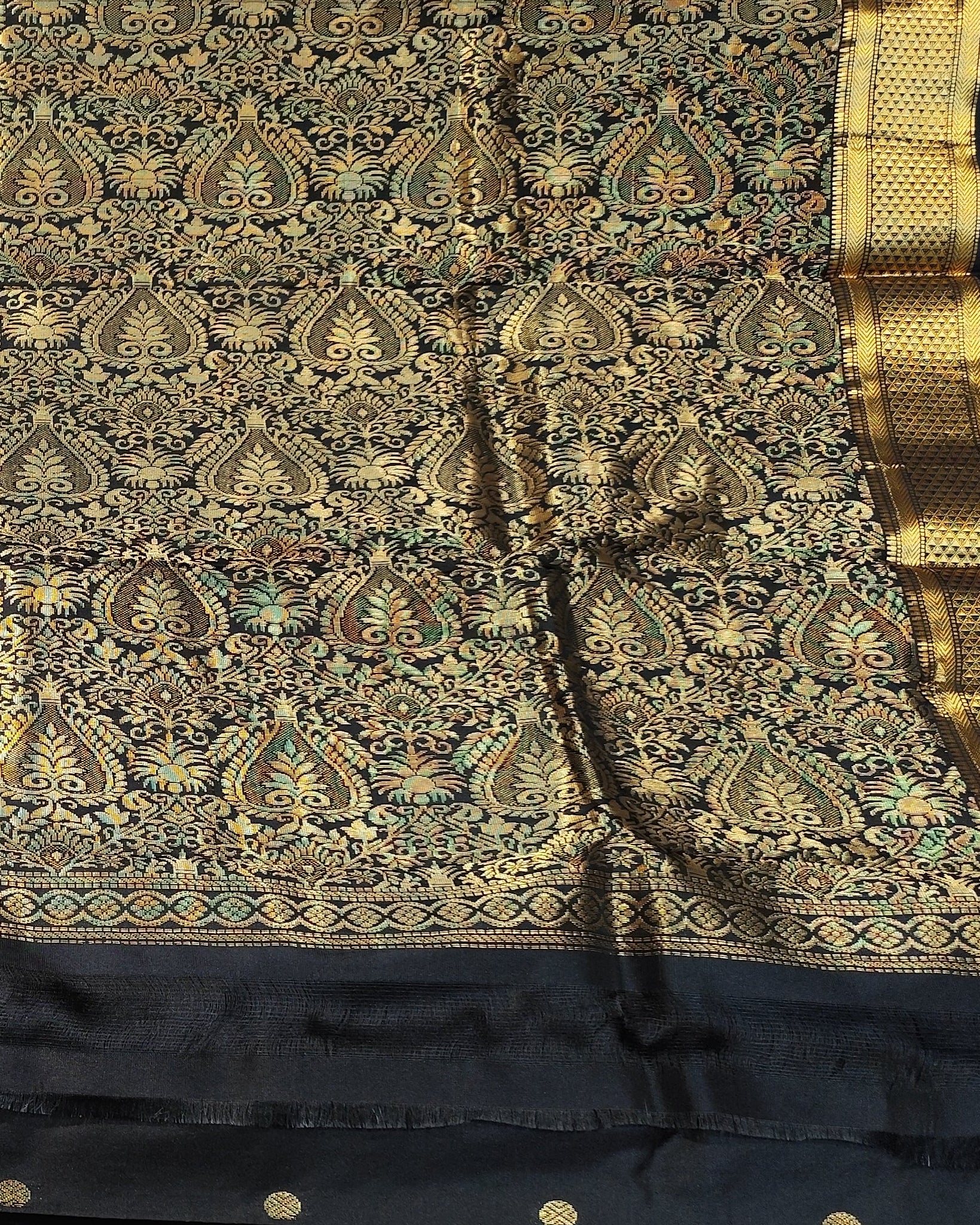 A black and gold Kanjeevaram silk. The saree features an intricate gold zari woven pattern on a black base, with a wide black border adorned with gold zari motifs and small gold buttons.