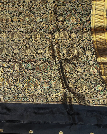 A black and gold Kanjeevaram silk. The saree features an intricate gold zari woven pattern on a black base, with a wide black border adorned with gold zari motifs and small gold buttons.