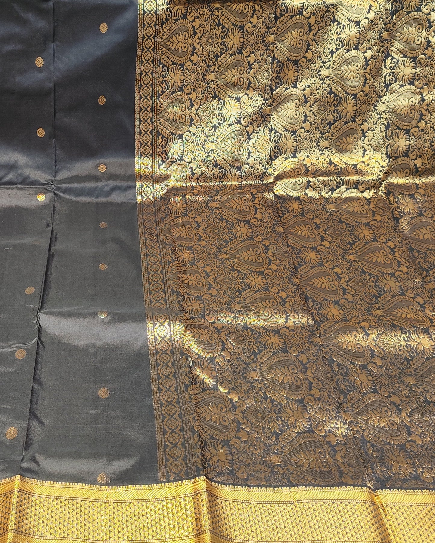 A black and gold Kanjeevaram silk saree displayed flat, showing its body, pallu, and ornate zari work border, with stacks of folded colorful sarees visible in the background.