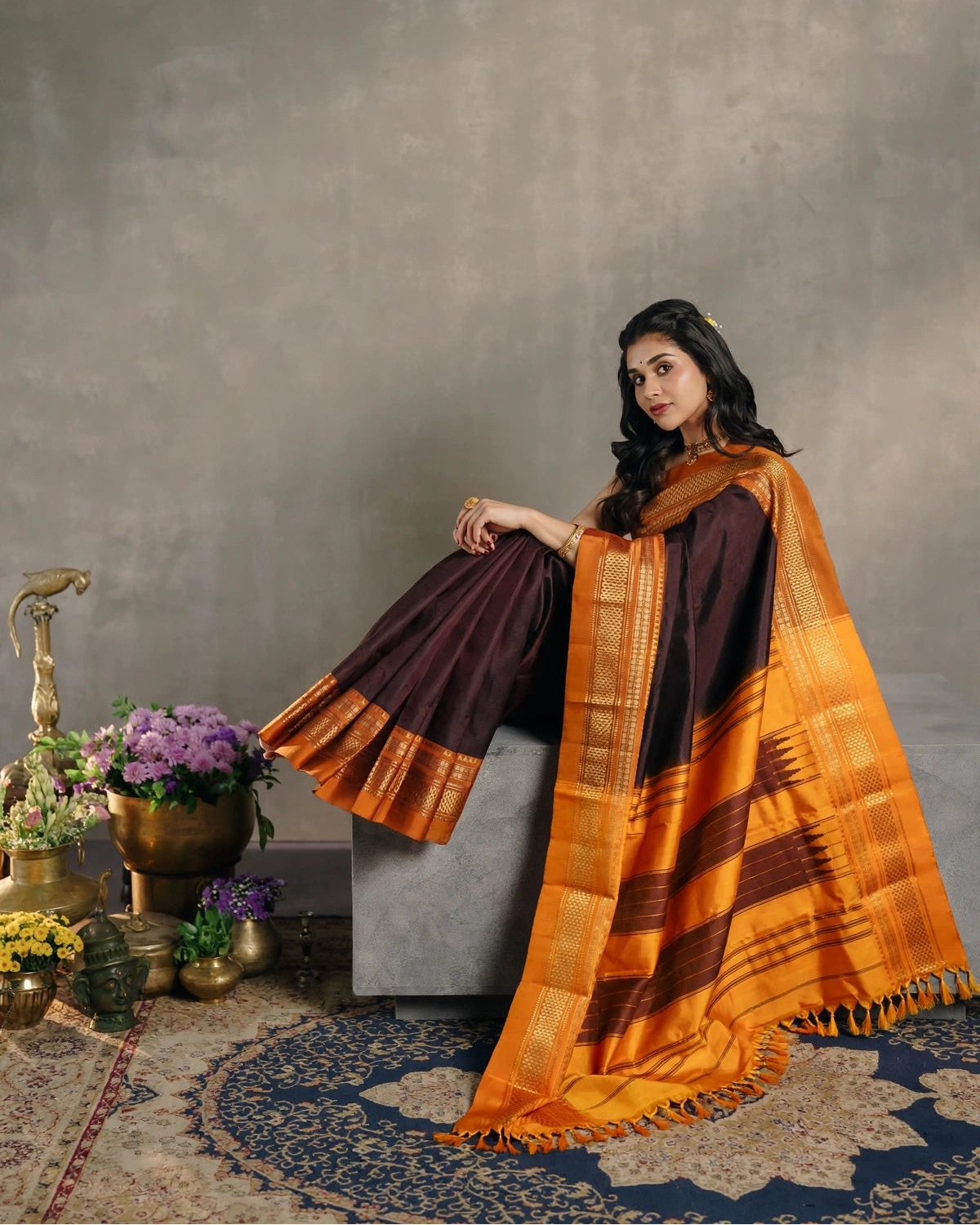 A woman models a dark brown and orange striped Ilkal Silk saree with a matching orange and brown checkered dupatta, seated on a gray stone block in a studio setting with decorative plants and a patterned rug.