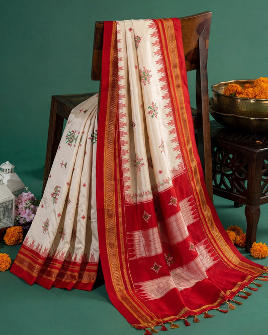 A cream and red Ilkal Temple motif silk saree draped over a dark wooden chair, decorated with intricate floral motifs and a red border with gold zari detailing, set against a dark green background with decorative marigold flowers.