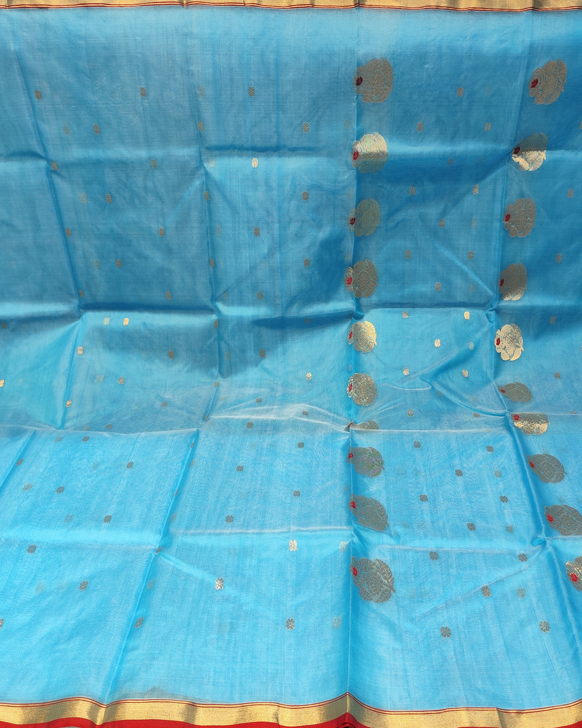 A light blue Chanderi saree or fabric drape displayed flat, featuring metallic silver circular motifs, small gold dots, and a red and gold border along the edge.
