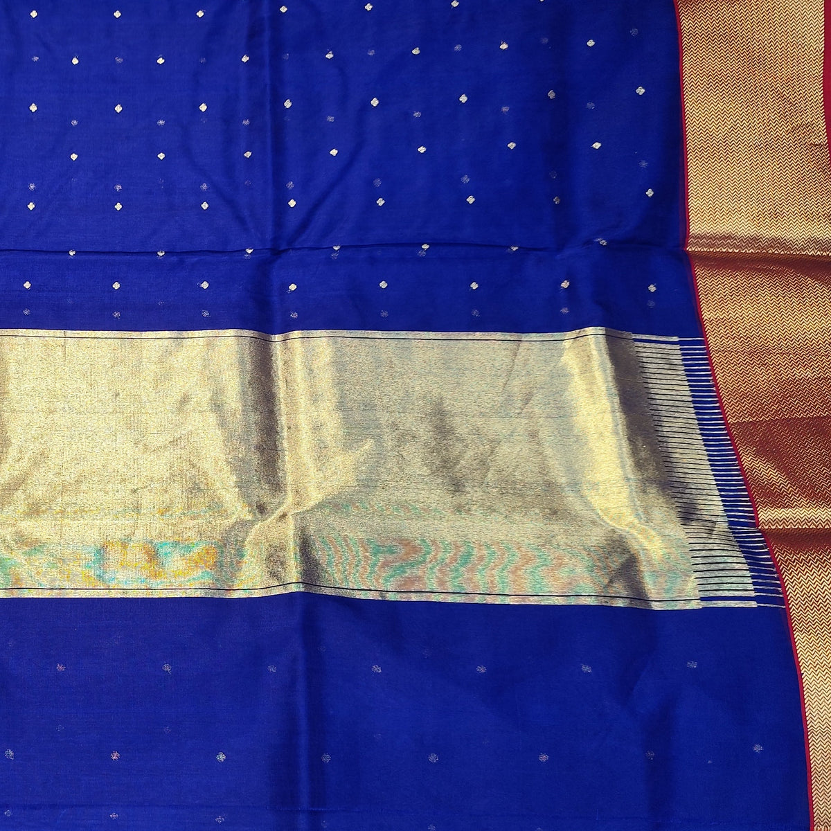 A royal blue Chanderi silk saree with a wide gold zari border and a central gold and teal patterned pallu, held up by a person in a fabric store.