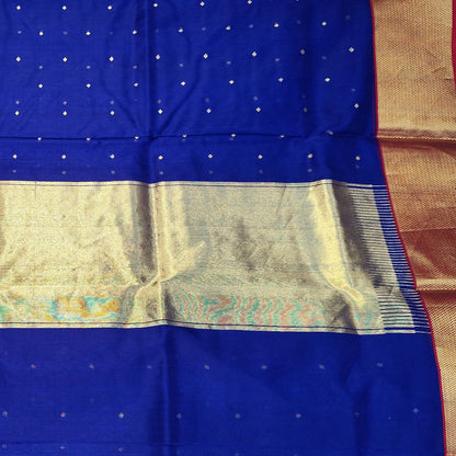 A royal blue Chanderi silk saree with a wide gold zari border and a central gold and teal patterned pallu, held up by a person in a fabric store.