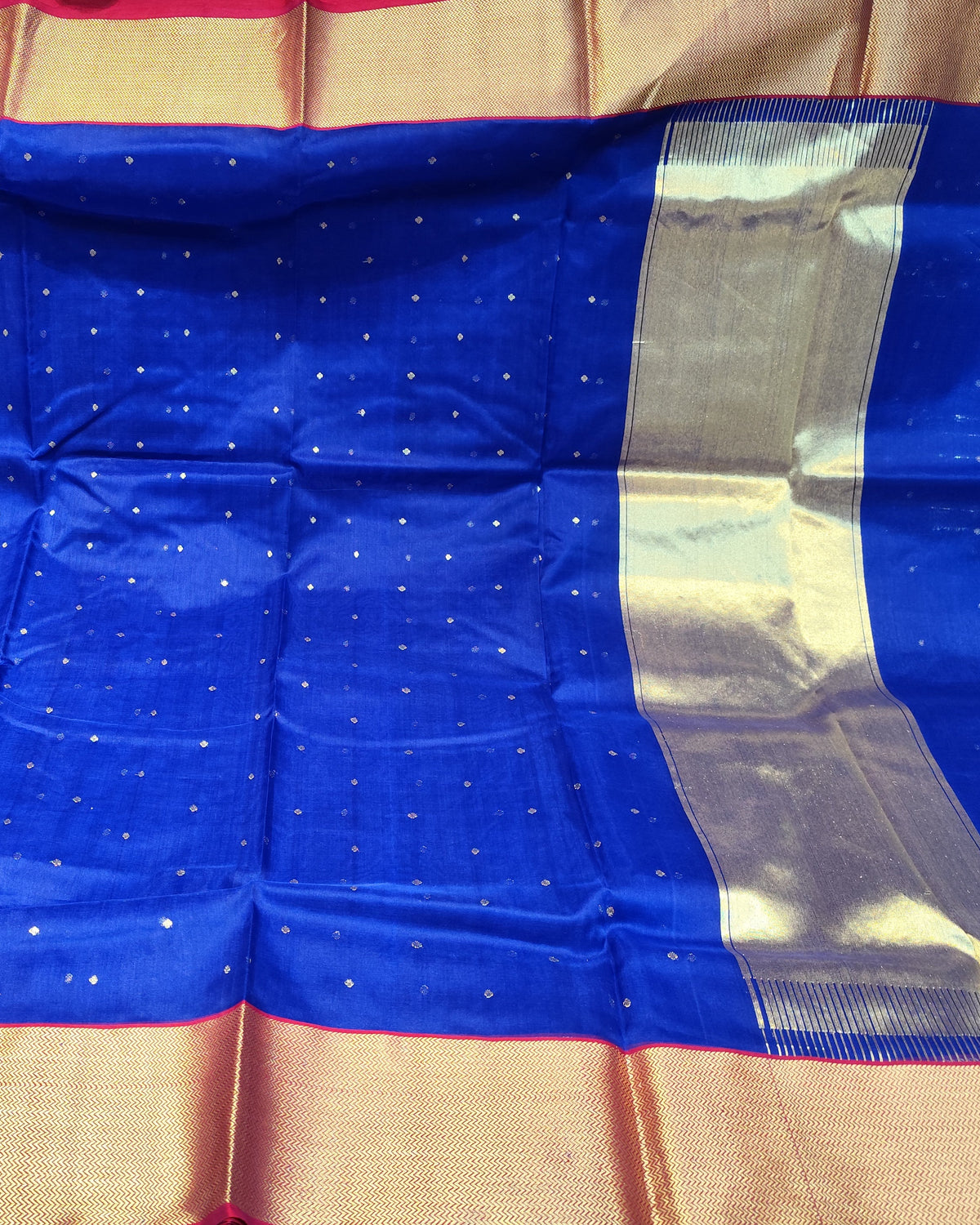A royal blue chanderi silk saree with small gold motifs and a wide gold zari border, displayed flat in what appears to be a fabric store.